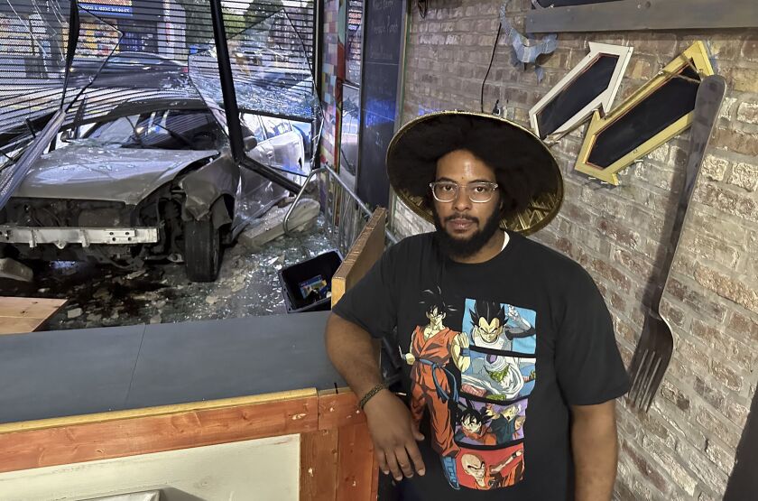 A man, Joe Black, stands in front of his and his wife, Tonya’s front window of their restaurant. The window has been damaged by a car that drove through their restaurant. The broken glass and a car sit in the window, with glass and other debris on the ground.